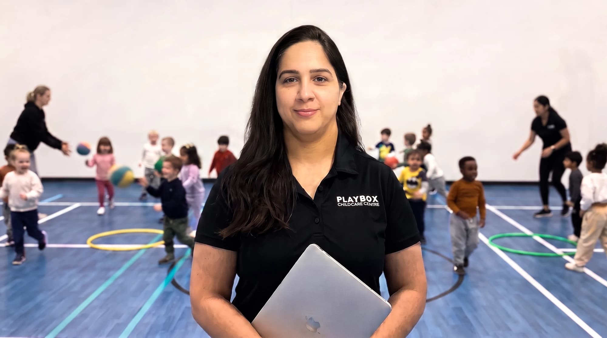 Miss Shalu, the Playbox manager, holding a laptop in the gymnasium while children play and two educators lead activities behind her